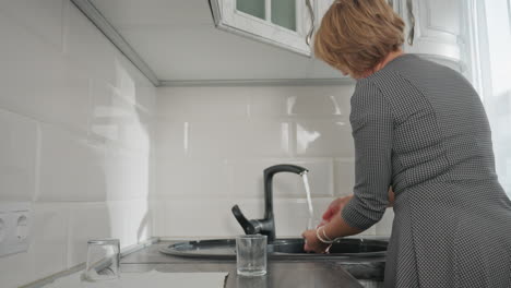 back view of young woman rinsing glass cup under running water at kitchen sink in bright clean kitchen environment with glasses placed on countertop showing focus on hygiene and domestic routine
