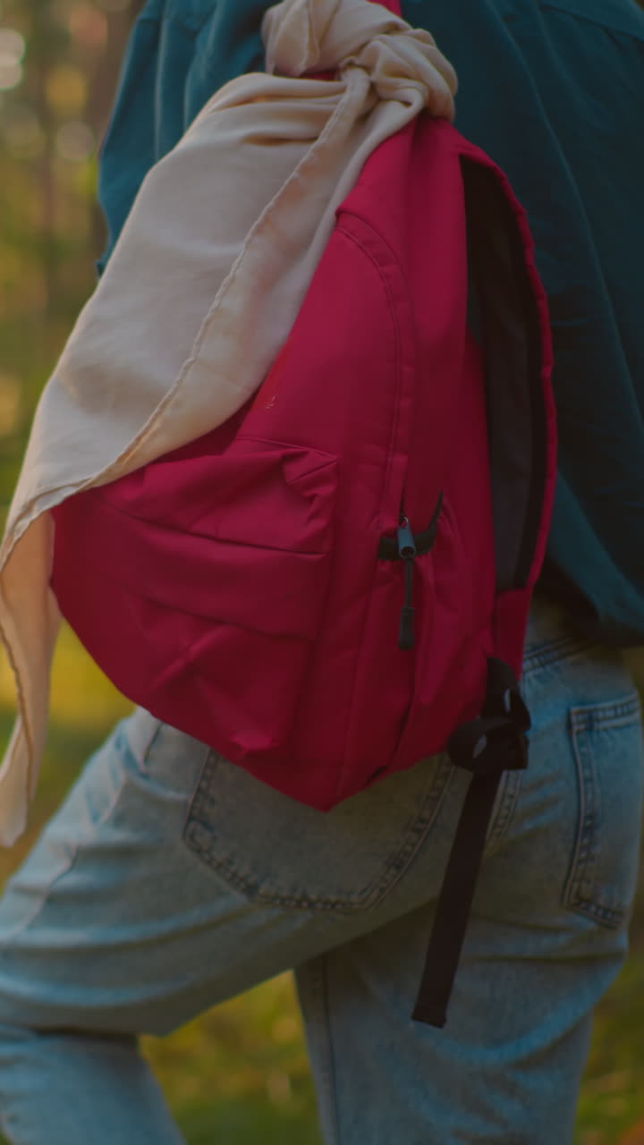 Back view of person wearing green shirt and blue jeans, with red bag and beige handkerchief draped over it, as golden sunlight creates a warm glow through forest trees