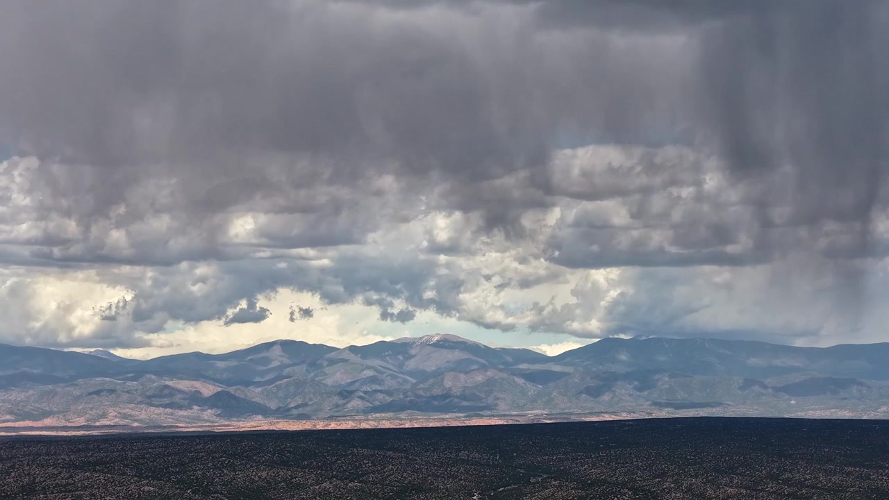 4K aerial timelapse captures close foreground rain. Cinematic motion reveals dramatic stacked cloud layers morphing and brewing over and behind a rugged southwest desert mountain peak.