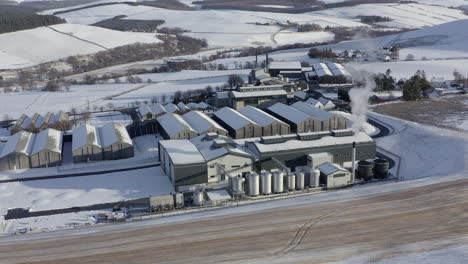 Aerial-view-of-the-Glenlivet-whisky-distillery-surrounded-by-snow-in-the-late-afternoon-sun-on-a-winters-day,-Moray,-Scotland---receding-shot