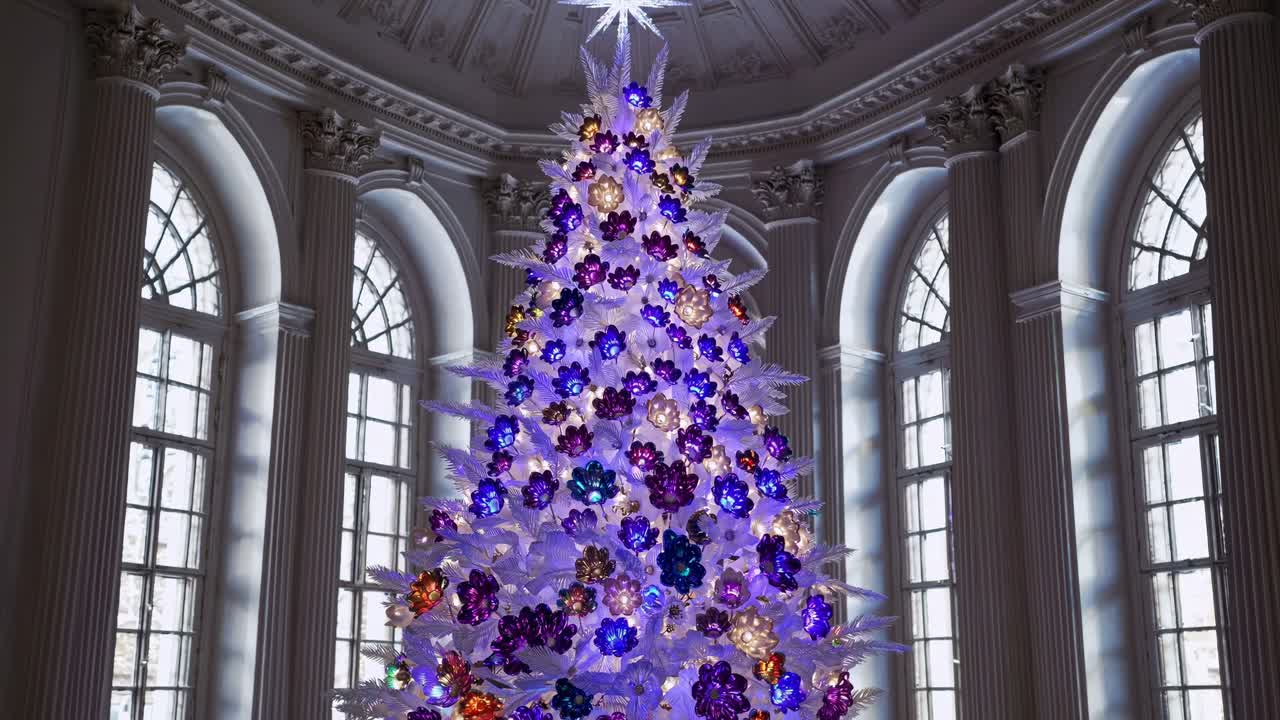 Christmas tree with white branches and colorful crystal flowers changing colors, placed in a luxurious palace hall with large windows and columns