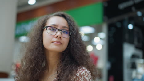 curious woman with glasses standing in busy mall, glancing around with interest, background features store displays, soft lighting, and people walking by