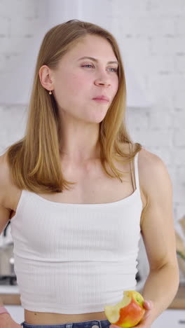 beautiful woman eating tasty apple indoors. young female with sporty body standing in the modern kitchen and eats fresh fruit. portrait of a lovely girl looking aside. vertical video