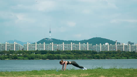 sporty woman stretches back and train core doing plank with knee tuck exercises on grassy lawn in han river park on summer morning, n seoul tower on namsan mountain in background