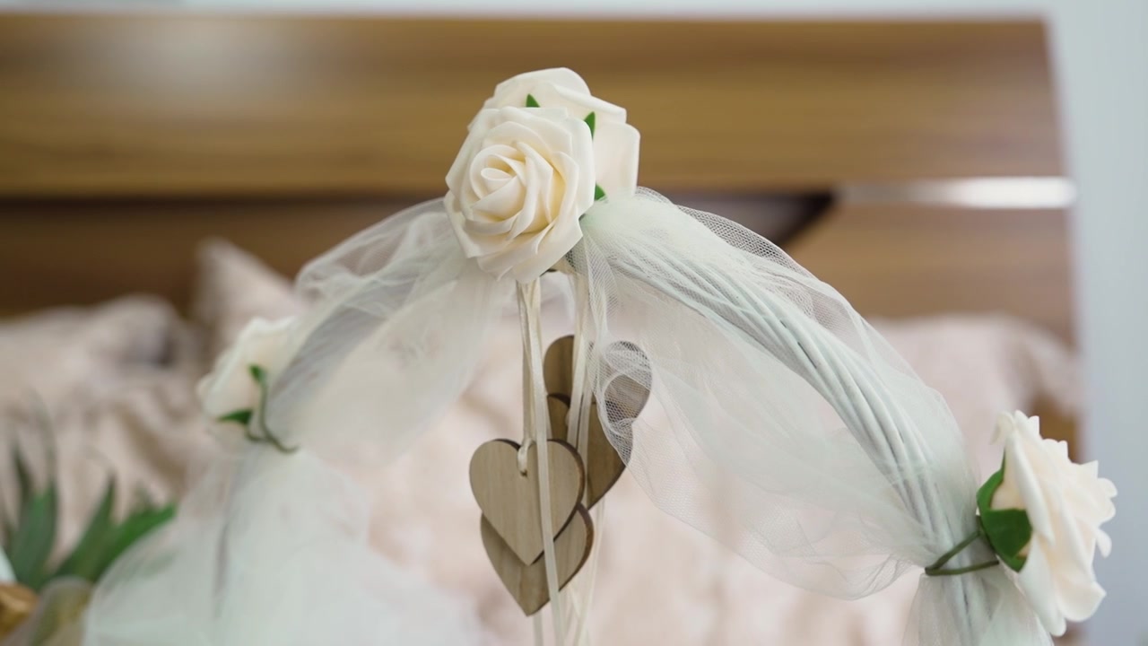 A simple decoration shows white flowers and wooden hearts hanging from a white frame. The scene is in a room with a bed in the background. The focus is on the wedding theme.