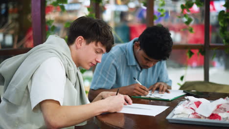 two close friends leaning over desk, intently focused on sketching on paper while ambient light highlights concentrated expressions and creative process unfolding in serene indoor setting