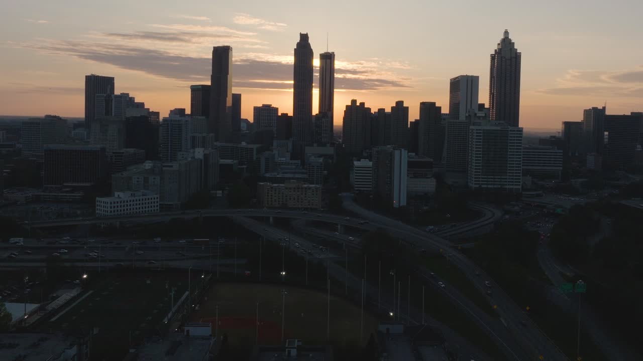 Static aerial shot of the downtown Atlanta cityscape skyline at sunset as the Atlanta skyscrapers are silhouetted and freeway traffic moves through the city.