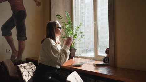 woman enjoys quiet moment sipping coffee near window as sunlight pours in, she gazes through window with warm smile, cozy interior features indoor plant, soft shadows, and artistic wall mural