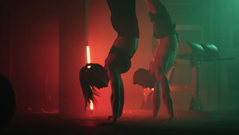 a man and a woman do a handstand in the gym together in slow motion. a strong man and a woman in a handstand walk across the gym floor in colored oil lamps