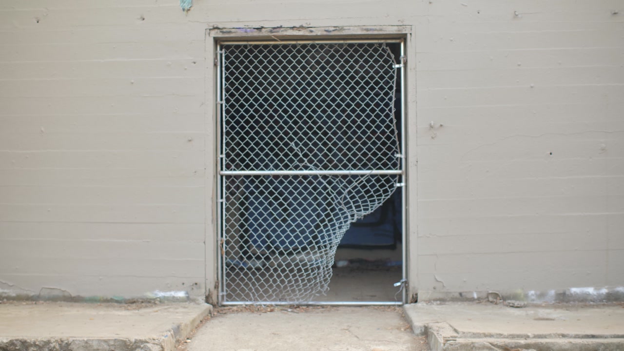A chain link fence with a hole in the bottom right corner leading to a graffiti laden abandoned room