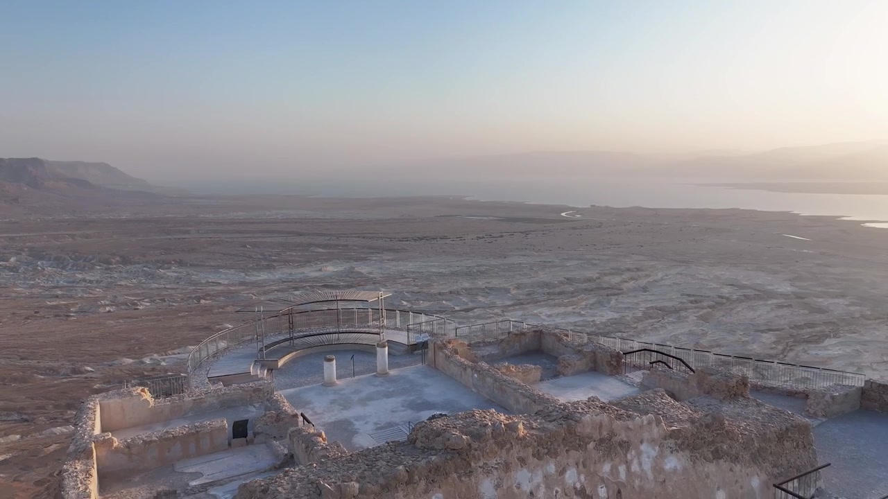 Drone tracking forward over the ancient stone walls and viewing point of Masada fortress revealing a wide panoramic view of the Judean desert landscape and Dead Sea during a hazy morning dawn.