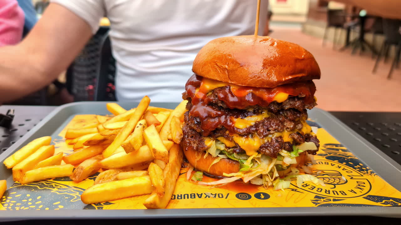 Four layer smash burger close up with fries on the side set on a tray on a food court outside, man with a white t shirt preparing to eat