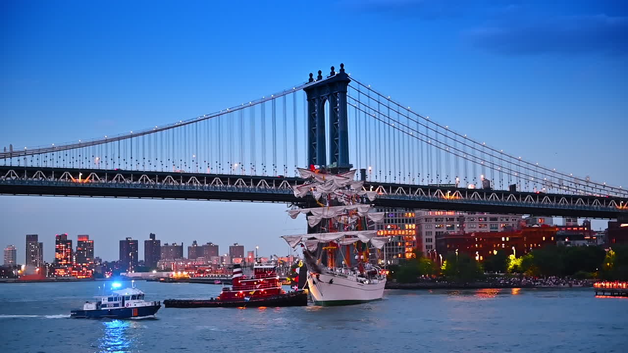 Un rimorchiatore e una motovedetta della polizia si avvicinano a una barca a vela a New York, USA. Il Manhattan Bridge con le luci accese al tramonto sullo sfondo