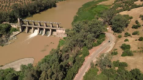 Vista-Aérea-De-Una-Represa-Hidroeléctrica-En-Un-Río-Y-Una-Carretera-Con-Un-Automóvil-Circulando-Por-Ella