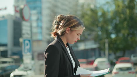 side view of bank manager in formal black suit walking outdoors while cross checking file in hand against urban background with parked cars, blurred street signs, and greenery on sunny day
