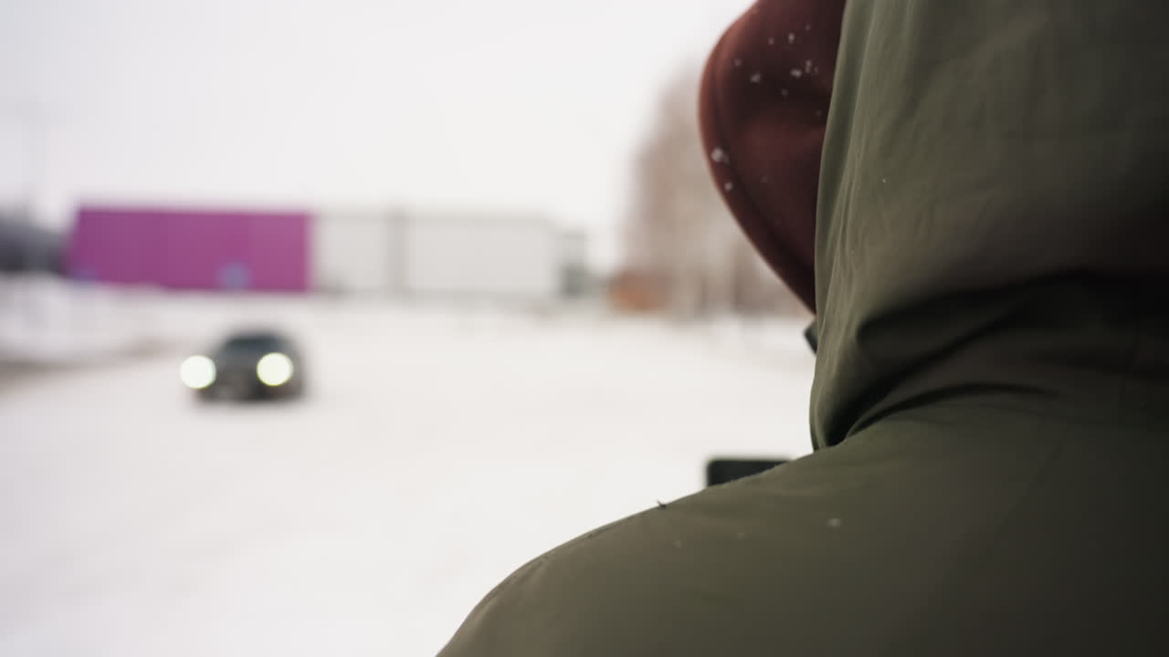 rear view of person in green hooded jacket standing outside in snow with blurred car drifting in distance, snowy road and buildings in background during cold winter day