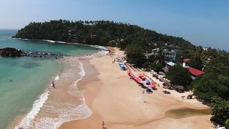 Nehmen-Sie-Eine-Aufnahme-Von-Mehreren-Bunten-Sonnenschirmen-Am-Breiten-Sandstrand,-Wo-Die-Menschen-Die-Sommerzeit-Genießen,-Sri-Lanka