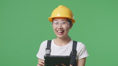 close up of asian woman worker wearing goggles and safety helmet using a tablet and pointing to side while standing in the green screen background studio