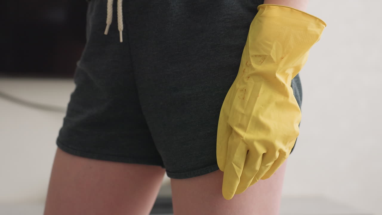 Close up of person in dark shorts and yellow cleaning gloves, standing indoors near wall and blurred television, capturing detail of hand gesture during household cleaning activity