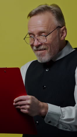 funny mature man with a folder isolated on yellow wall. happy old man in glasses writes on paper and depicts funny face. positive emotions. vertical video