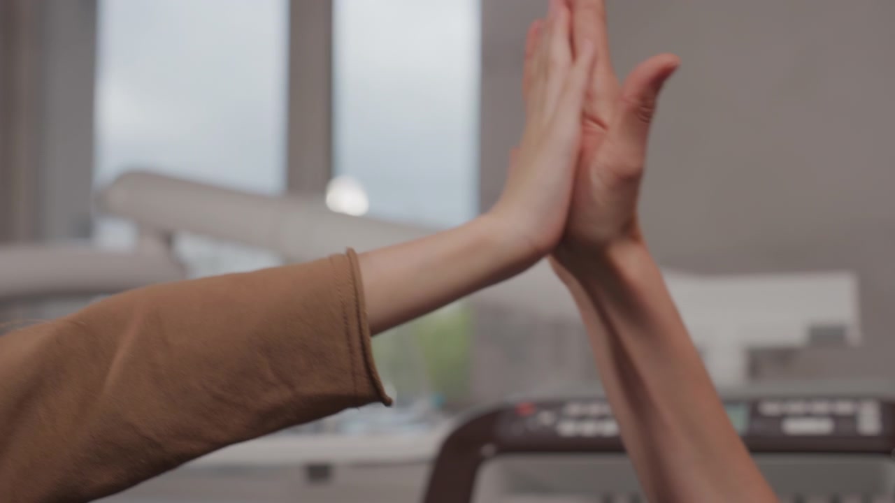 Primo piano di due mani che si protendono l'una verso l'altra in uno studio dentistico. Una mano indossa una manica marrone mentre lo sfondo mostra attrezzature mediche sfocate e grandi finestre con luce diurna.