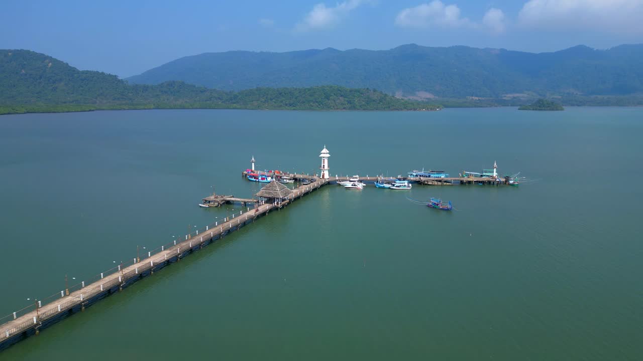 white lighthouse, long way pier extending into the bay with numerous boats docked and tropical islands in the background. Majestic aerial view panorama overview static tripod hovering drone