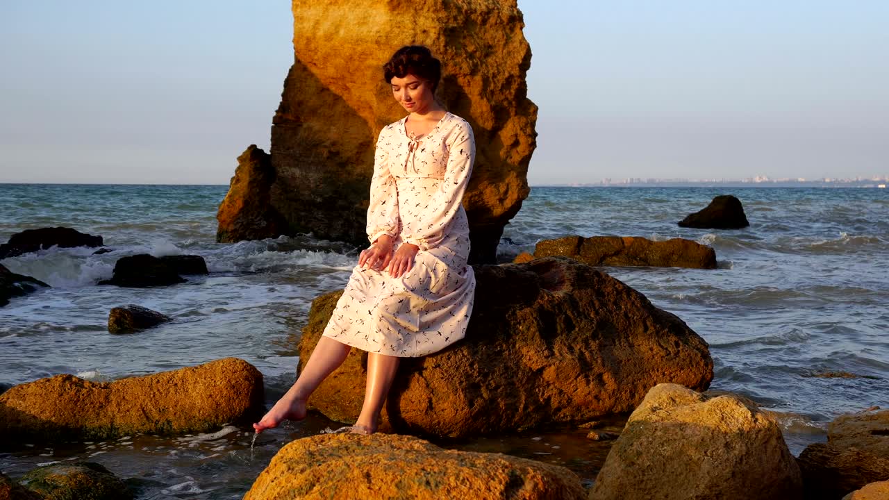 очаровательная белая брюнетка в светло-розовом платье сидит на скале в море во время восхода солнца