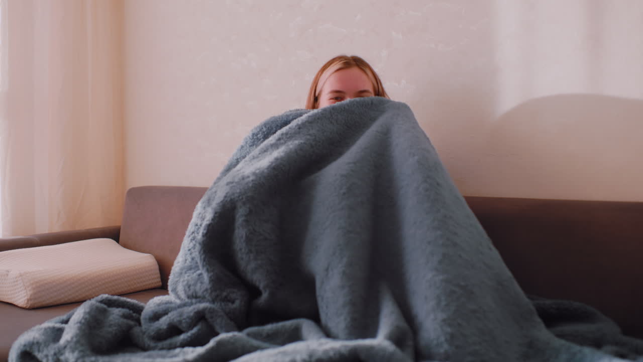 Playful woman sitting on sofa covering herself with fluffy blanket while smiling and looking down in soft morning light near curtain, creating cozy indoor moment with cheerful relaxed energy