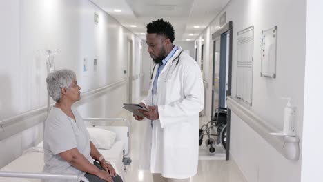 diverse serious male doctor with tablet talking to senior patient on bed in slow motion, unaltered