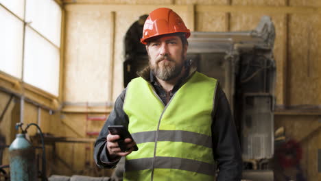 Trabajador-Con-Chaleco-Y-Casco-De-Seguridad-Sosteniendo-Un-Teléfono-Inteligente-Y-Mirando-La-Cámara-En-Un-Parque-Logístico