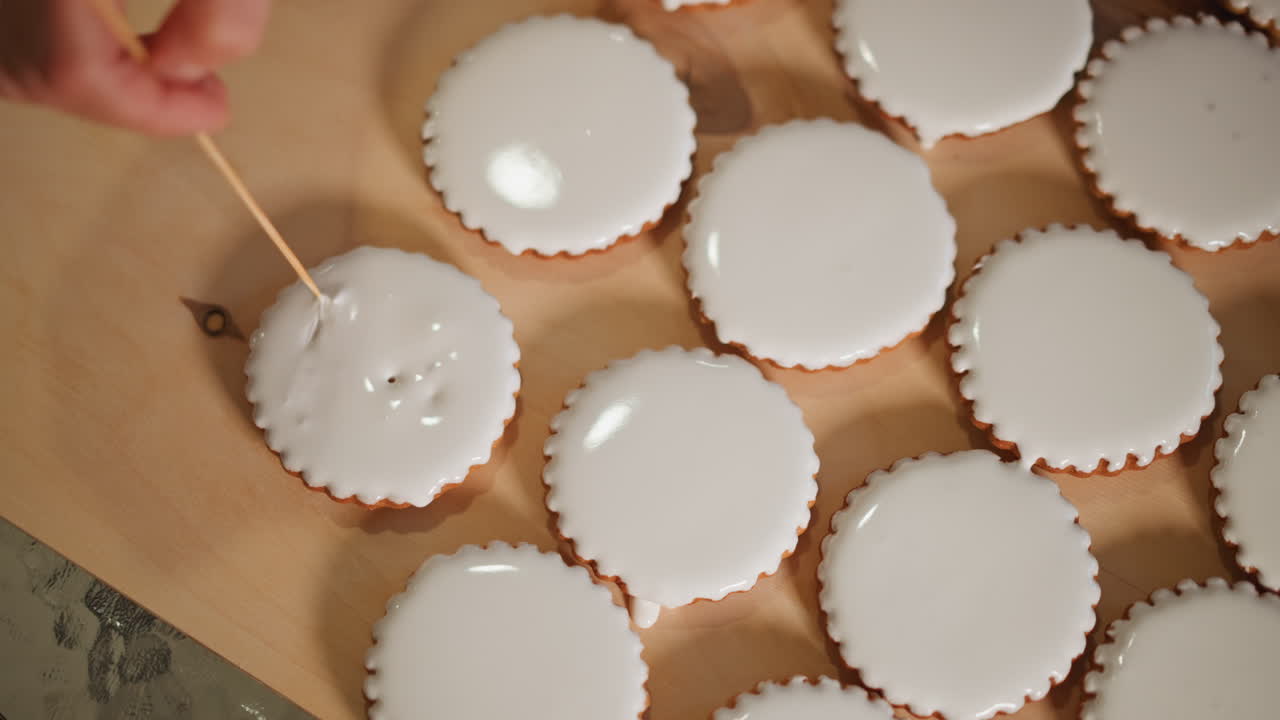 Hand testing iced mini pies for texture and glaze consistency with toothpick, inspection across batch of tartlets on countertop tray, glossy white icing, studio lighting, production vibe