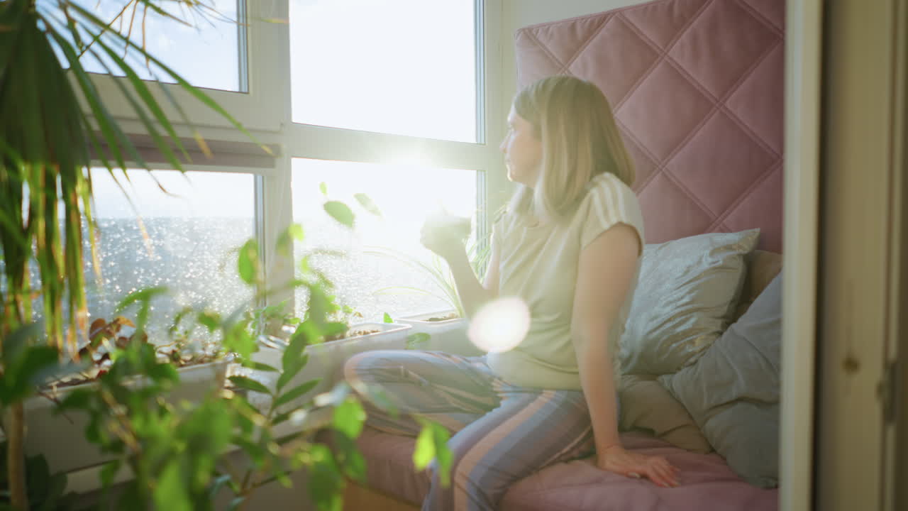 Woman in casual clothes sits on cozy window seat with striped pants, drinking hot beverage from glass cup, surrounded by green plants and sunlight, enjoying calm relaxation during peaceful morning at home