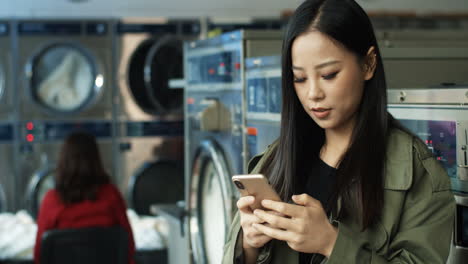 pretty woman with long dark hair tapping and texting message on smarphone while standing in laundry service room