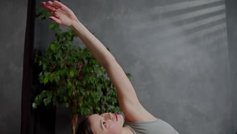 confident and calm athletic brunette girl in a gray sports top and green pants does yoga on the red sports carpet and stretches her arm up during her warm-up and stretching at home while doing sports