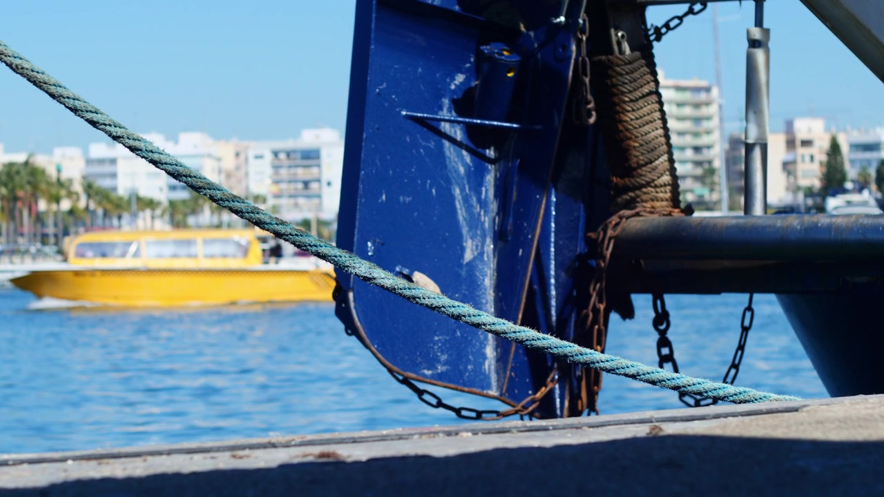 Poppa di un peschereccio nel porto di Santa Pola, Spagna, con un taxi acqueo giallo sfocato che naviga verso l'isola di Tabarca sullo sfondo. Scatto cinematografico.