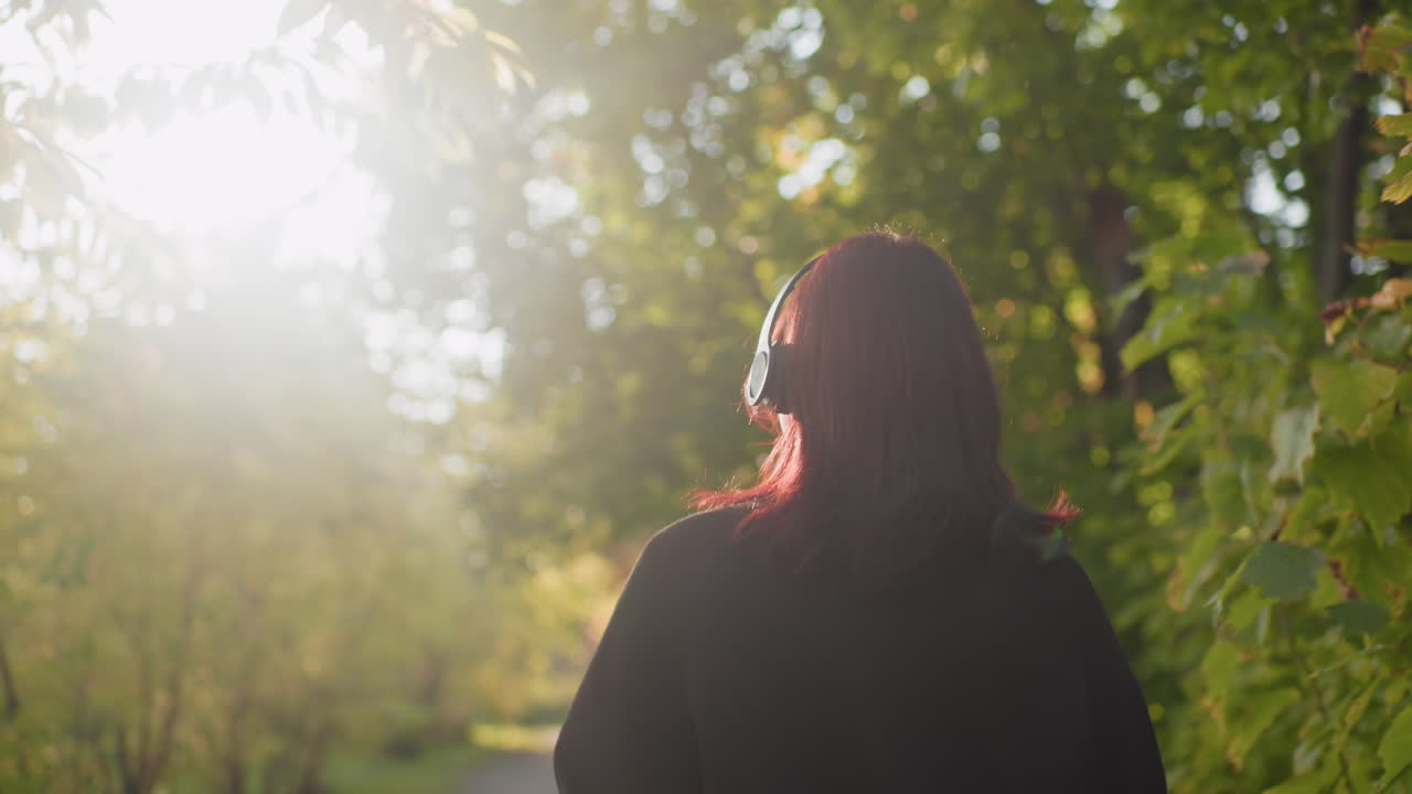 Medium back view of nature lover in headphones strolling through sunlit arboretum path, soaking in music, relaxed pace, warm glow, green leaves around, carefree mood, casual coat, soft breeze