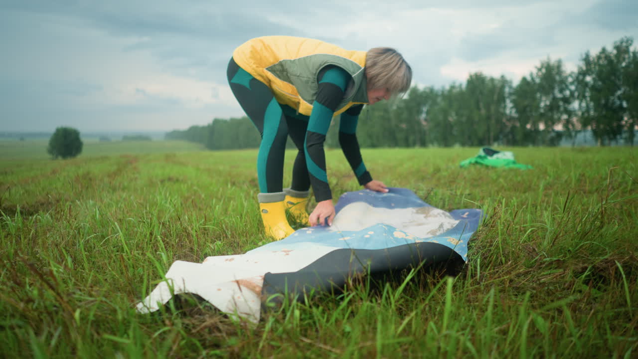 Middle-aged woman adjusting her colorful mat on grassy field with green bag in the background, she sets up in a vast open field, with trees in the distance under a cloudy sky
