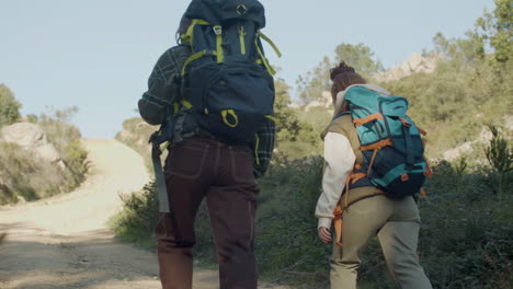 vista trasera de dos jóvenes excursionistas caminando por un camino de tierra en un día soleado mientras llevan mochilas y hablan 1