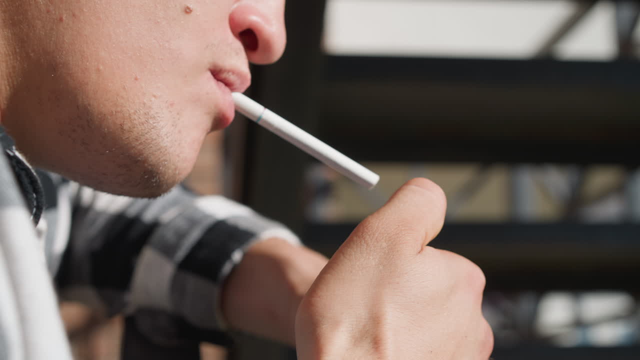 Close up of man holding cigarette in mouth while igniting lighter to smoke, with focus on fingers and mouth in warm sunlight, featuring soft blurred industrial urban background