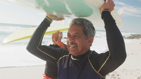 Glückliches-älteres-Hispanisches-Paar,-Das-Mit-Surfbrettern-über-Dem-Kopf-Am-Strand-Spaziert