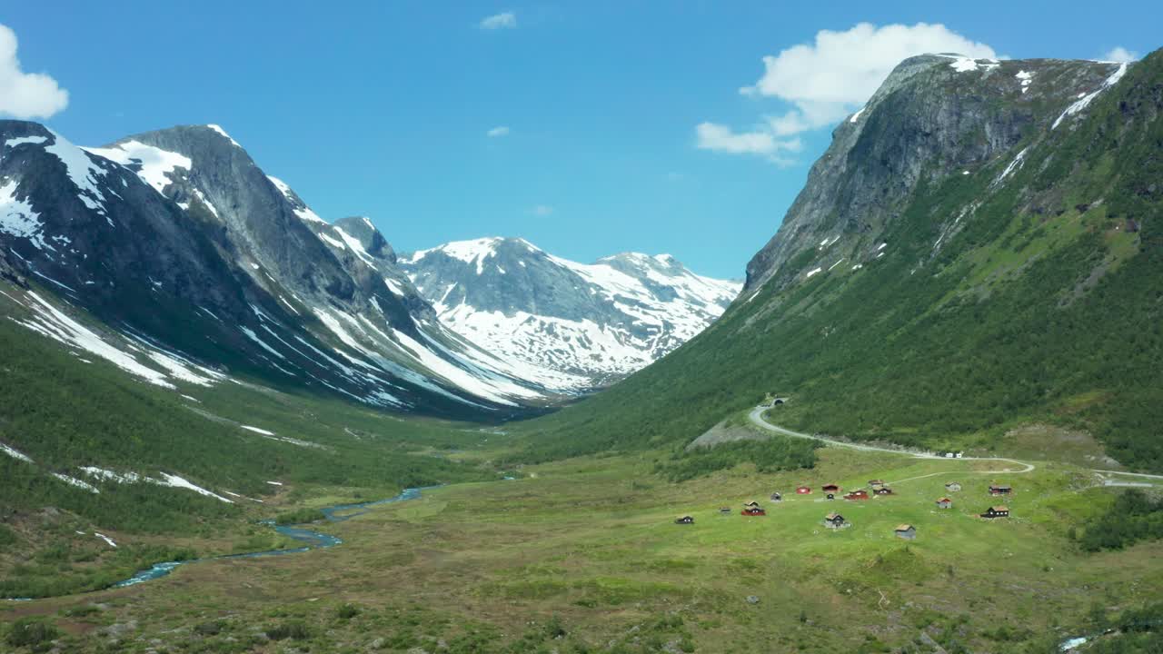 Aerial view of a vast green valley surrounded by towering mountains in Norway and cabin community in lowlands, with patches of snow on the peaks under a clear blue sky