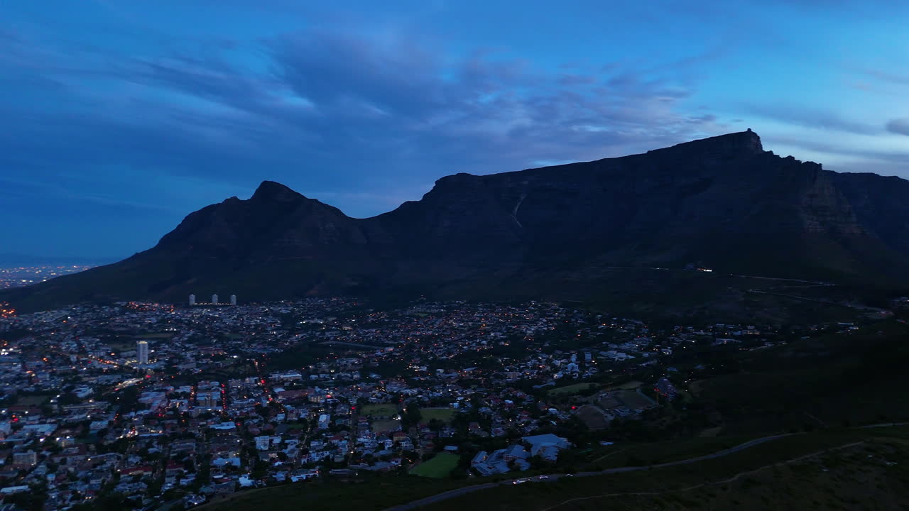 Cinematic aerial view of Table Mountain in Cape Town, South Africa, dramatic rocky cliffs with coastal cityscape at sunset, clouds rolling over peaks and golden evening light