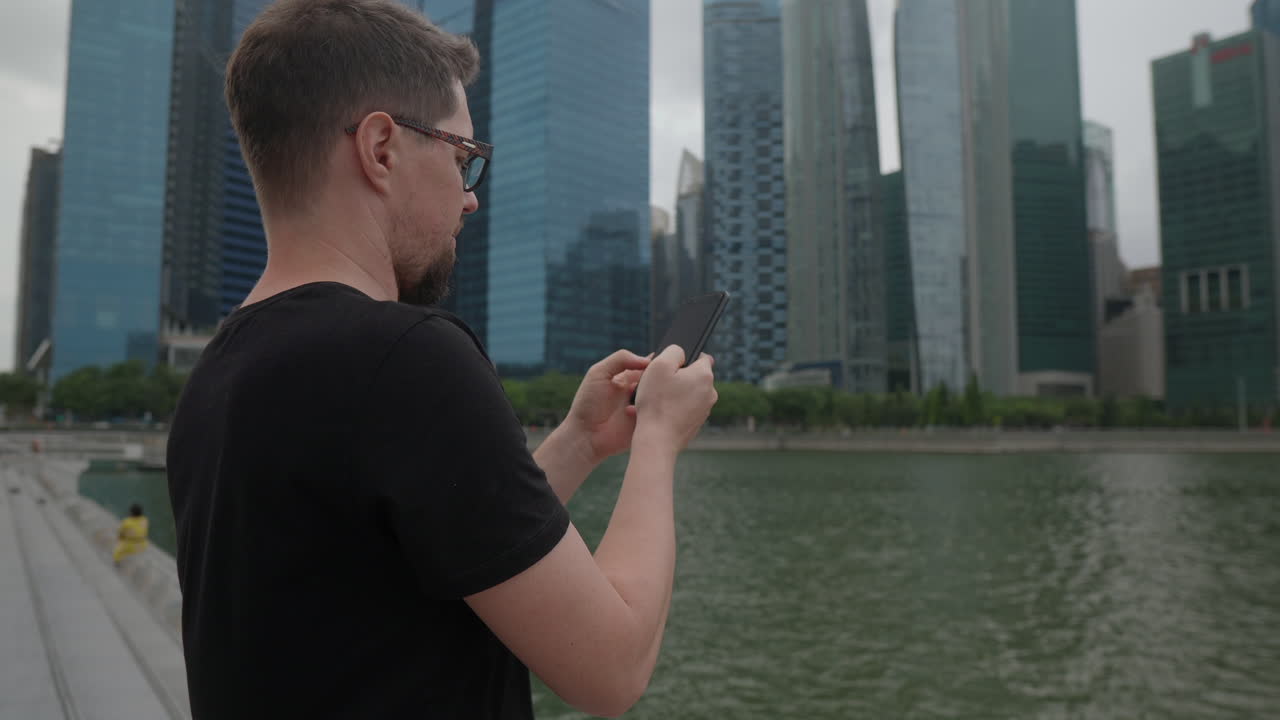 Man using smartphone by a river in a city with skyscrapers