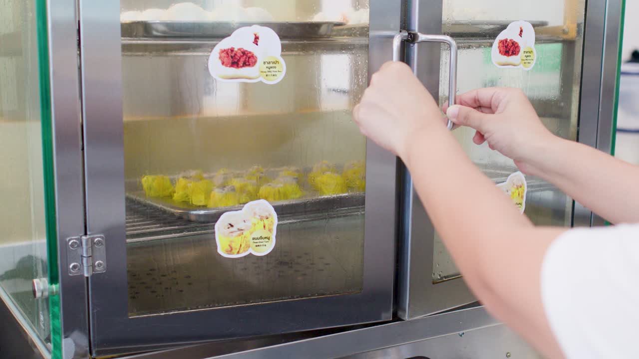 Una persona chiude gli sportelli di vetro di una vaporiera alimentare professionale contenente vassoi di ravioli.