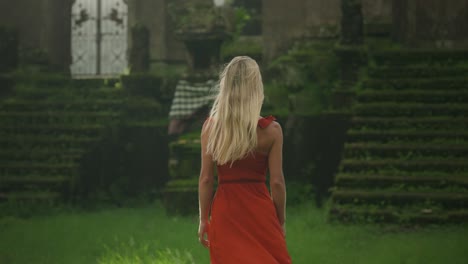 Misteriosa-Mujer-Rubia-Vestida-De-Rojo-Caminando-Sobre-El-Campo-De-Hierba-Hacia-Los-Escalones-De-Piedra-Del-Antiguo-Templo,-Escena-De-Fantasía