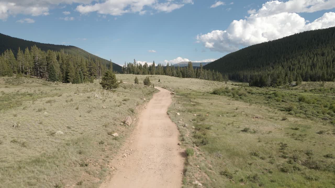 Aerial view following a remote dirt road near Guanella Pass, Colorado. Dolly movement with green grass, forest and clouds in the distance.