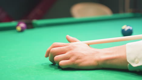 primer plano de la mano descansando en la mesa de billar verde con palo de taco posicionado para jugar. la manga blanca nítida contrasta con el fieltro verde vibrante. el fondo borroso con la pelota de billar agrega profundidad y juego