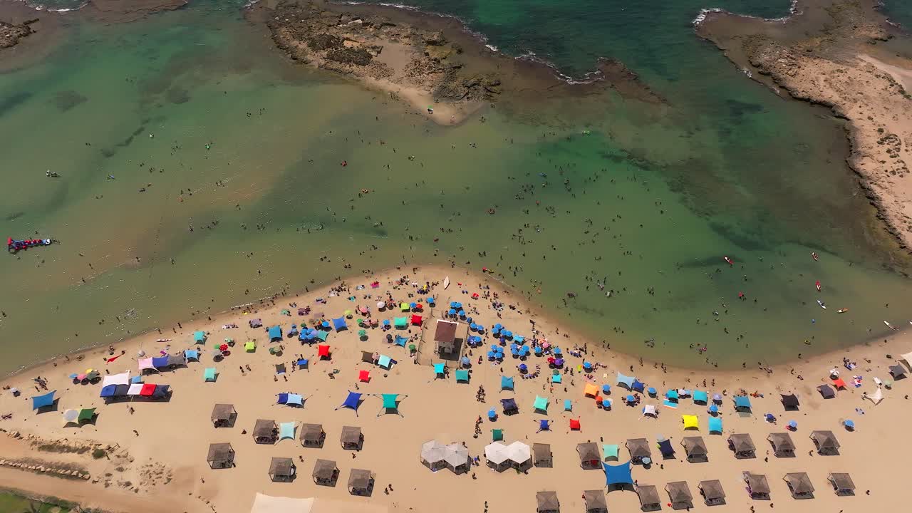 Dia de verão na praia com pessoas na água e sob guarda-sóis coloridos, filmagem de drone de cima para baixo.