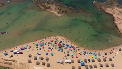 Día-De-Verano-En-La-Playa-Con-Gente-En-El-Agua-Y-Bajo-Sombrillas-Coloridas,-Imágenes-De-Drones-De-Arriba-Hacia-Abajo