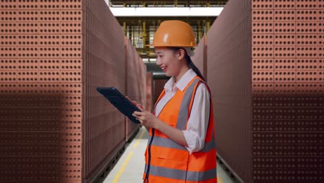 Side View Of Asian Female Engineer With Safety Helmet Looking At The Tablet In Her Hand And Looking Around While Standing With Red Brick Packed in Stacks Are Stored